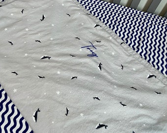 May include: A gray crib sheet with a white background and a navy blue and white wave pattern border. The sheet has a repeating pattern of small white stars and black whales. The sheet is personalized with the name "Troy" embroidered in blue thread.