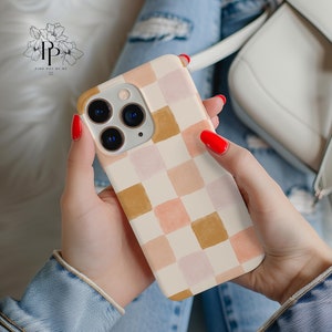 May include: A phone case with a cream-colored background and a checkerboard pattern in shades of peach, pink, and brown. The phone case has a gold-colored camera ring and is held in a person's hands.