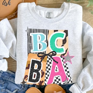 May include: A light gray sweatshirt with the letters "BCBA" in a colorful design. The letters are in various colors, including blue, green, black, and pink, with a black bow and star accents. The sweatshirt is folded on a wooden surface.