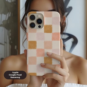 May include: A phone case with a checkered pattern in shades of peach, tan, and cream. The case is held up in front of a person's face. The text on the case reads "Apple, Google Pixel, Samsung."