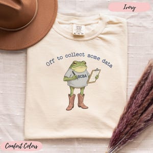 May include: Ivory Comfort Colors t-shirt featuring a frog graphic. The frog is wearing a grey shirt with "BCBA" on it, cowboy boots, and holding a clipboard. The text "Off to collect some data" is above the frog.