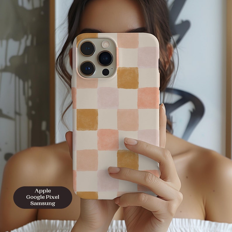 May include: A phone case with a checkered pattern in shades of peach, tan, and cream. The case is held up in front of a person's face. The text on the case reads "Apple, Google Pixel, Samsung."