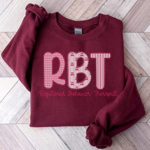 May include: Burgundy sweatshirt with the letters "RBT" in pink, featuring gingham, stripes, and cherry accents. The words "Registered Behavior Therapist" are printed below the letters. The sweatshirt is folded on a light wood surface.