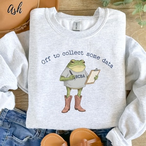May include: Pale grey sweatshirt with the text "Off to collect some data." The graphic shows a cartoon frog wearing a shirt that says "BCBA" and cowboy boots, holding a clipboard and pen.