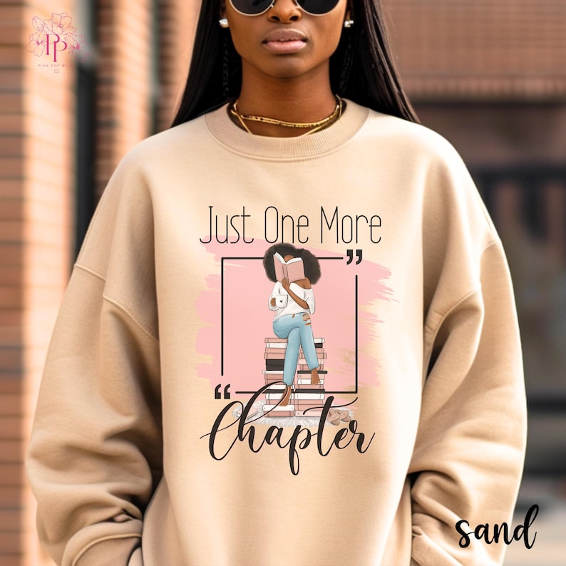 May include: Sand-colored sweatshirt with the text "Just One More Chapter" and a graphic of a person reading a book. The graphic shows a person sitting on a stack of books, reading. The word "sand" is printed at the bottom right.