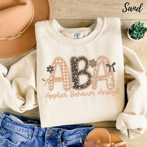 May include: A cream-coloured sweatshirt featuring the letters "ABA" in a decorative font. The letters are embellished with floral and patterned designs. The words "Applied Behavior Analyst" are printed below the letters in a script font.
