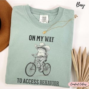 May include: A pale green t-shirt featuring the text "ON MY WAY TO ACCESS BEHAVIOR" and a graphic of a frog in a cowboy hat riding a bicycle. A gold necklace is draped across the shirt.