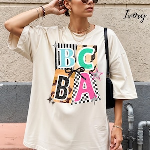 May include: Ivory-colored oversized t-shirt with a colorful graphic design. The graphic features the letters "BCBA" in various colors and patterns, including black and white stripes, a checkered pattern, and a leopard print.