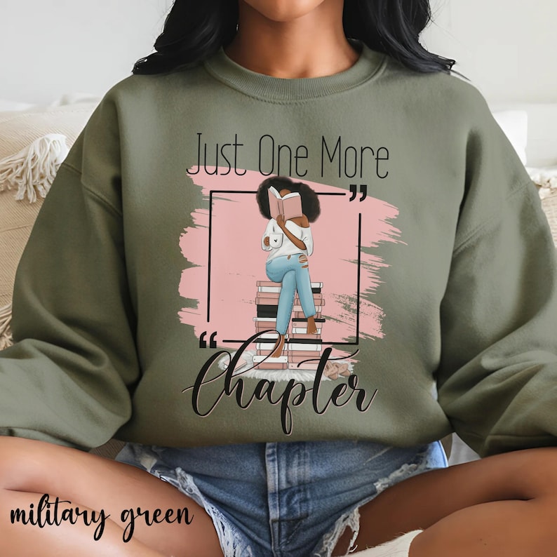 May include: Military green sweatshirt with the text "Just One More Chapter" and a graphic of a person reading a book. The graphic is set against a pink background. The sweatshirt is a solid color.