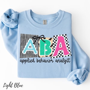 May include: Light blue sweatshirt with the letters "ABA" in a colorful design. The "A" is light blue, the "B" is green, and the second "A" is pink. The words "applied behavior analyst" are printed below the letters.