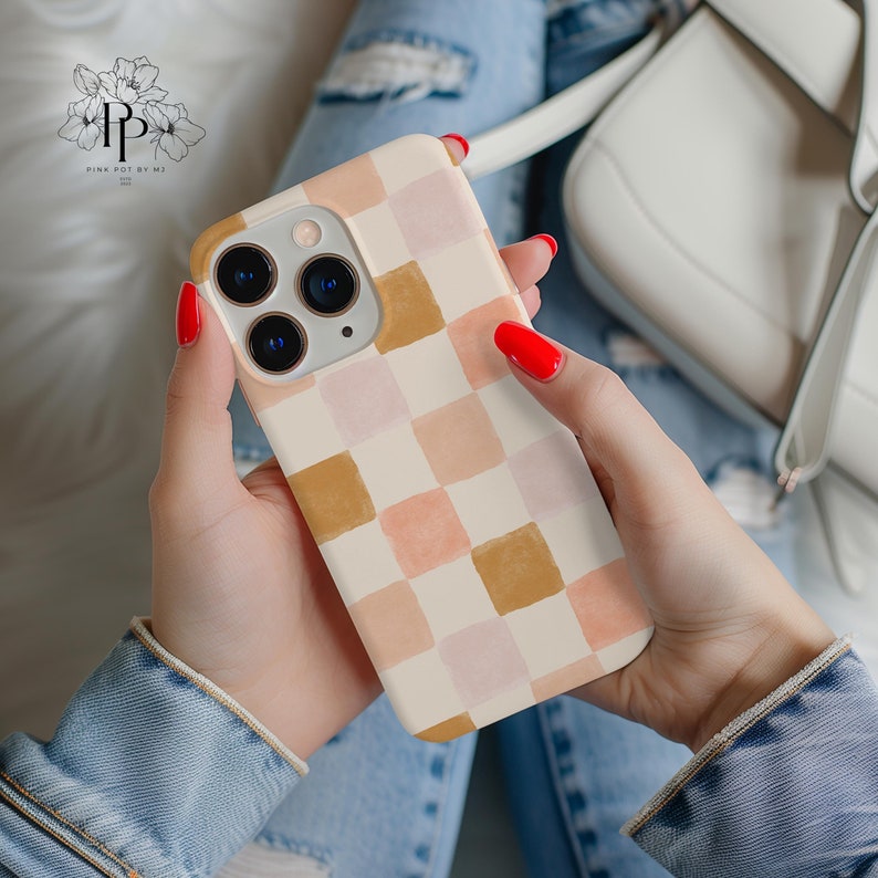 May include: A phone case with a cream-colored background and a checkerboard pattern in shades of peach, pink, and brown. The phone case has a gold-colored camera ring and is held in a person's hands.