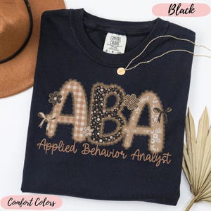 May include: Black Comfort Colors t-shirt featuring the letters "ABA" in a decorative design. The letters are in a mix of patterns, including floral and plaid. The words "Applied Behavior Analyst" are printed below.