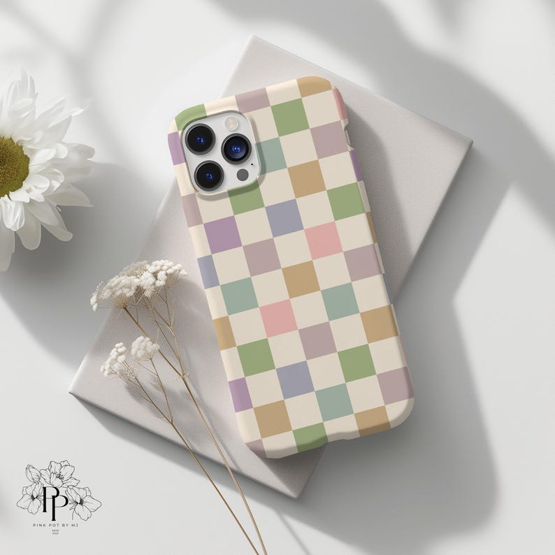 Puede incluir: Una funda de tel&eacute;fono de colores pastel con un patr&oacute;n de tablero de ajedrez en tonos verdes, morados, rosas y beige. La funda est&aacute; sobre una superficie blanca con una flor blanca y delicadas ramitas de flores secas. La funda tiene un recorte para la c&aacute;mara.