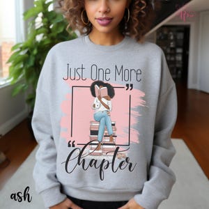 May include: Ash gray sweatshirt with the text "Just One More Chapter" and an illustration of a person reading a book. The graphic includes a pink background and a stack of books. The word "ash" is printed at the bottom left.