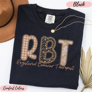 May include: Black Comfort Colors t-shirt with the letters "RBT" in a decorative design. The "R" is a tan and white gingham pattern, the "B" is a floral pattern, and the "T" is a floral pattern. The words "Registered Behavior Therapist" are below the letters.
