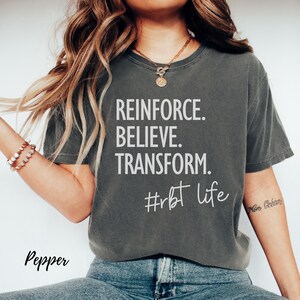 May include: Dark gray t-shirt with the words "REINFORCE. BELIEVE. TRANSFORM." in white, along with the hashtag "#rbt life." The word "Pepper" is written in cursive at the bottom left.
