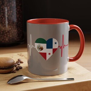 May include: A gray ceramic coffee mug with a red interior and handle. The mug features a heart design with the flags of Mexico and Panama, along with a heartbeat graphic. A spoon and a macaron are on a wooden surface.