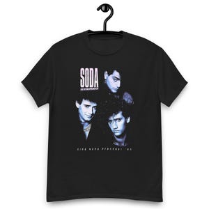 May include: A black short-sleeve t-shirt featuring a blue-toned graphic print of three individuals. The text SODA STEREO is printed in white above the figures, and the text GIRA NADA PERSONAL '85 is printed in white below the figures on the chest.