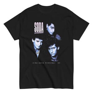 May include: A black short-sleeve t-shirt featuring a blue-toned graphic print of three individuals. The text SODA STEREO is printed in white above the figures, and the text GIRA NADA PERSONAL '85 is printed in white below the figures on the chest.