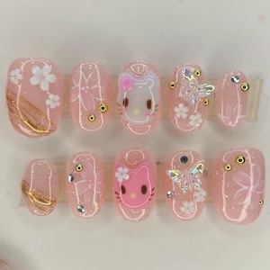 May include: A set of ten pink press-on nails. Each nail features a different design, including floral patterns, gold accents, and a Hello Kitty character. Some nails have small rhinestones and butterfly embellishments.