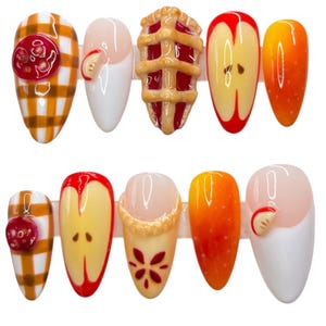 May include: A set of ten artificial nails with various designs. The designs include a red button on a yellow plaid background, a white nail with a small apple slice, a miniature pie, and apple slice designs. The nails are in various shades of red, yellow, and orange.