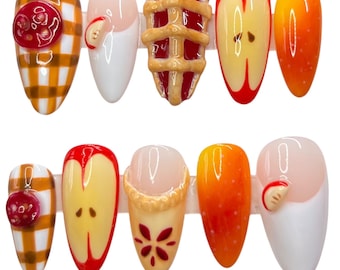Peut inclure: Dix faux ongles décorés de motifs variés. Les motifs comprennent un bouton rouge sur fond jaune à carreaux, un ongle blanc avec une petite tranche de pomme, une tarte miniature et des motifs de tranches de pomme. Les ongles sont dans des tons de rouge, jaune et orange.