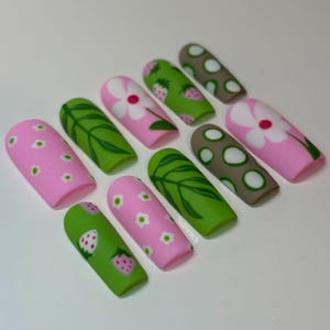 May include: A set of ten press-on nails featuring a matte finish with various designs including pink with small white flowers, green with leaf patterns, pink with large white flowers, green with pink strawberries, and brown with white circles.