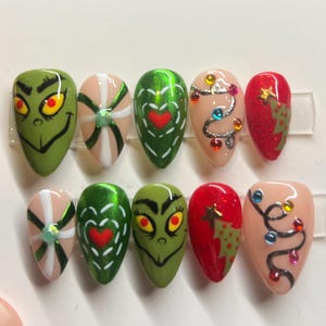 May include: A set of ten almond-shaped, painted artificial nails. Designs include a Grinch face, a heart, a Christmas tree, and a string of lights. Colours include green, red, white, and nude.