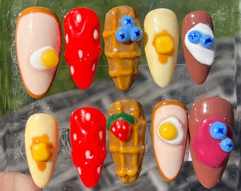 Peut inclure: Ensemble de dix faux ongles avec différents motifs de nourriture pour le petit-déjeuner. Les motifs comprennent un œuf au plat, une fraise, une gaufre, du sirop et des myrtilles.