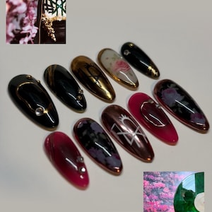 May include: A set of ten almond-shaped press-on nails featuring various designs including glossy black with gold accents, metallic gold with black symbols, floral patterns, deep translucent red, and silver embellishments like small rhinestones.