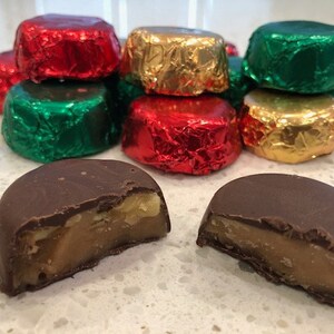 Pode incluir: Um close-up de dois doces de caramelo cobertos de chocolate, cortados ao meio para revelar o recheio de caramelo. Os doces são cercados por uma variedade de chocolates embrulhados em papel alumínio vermelho, verde e dourado.