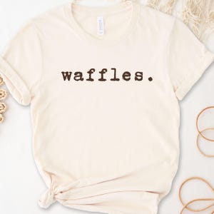 May include: A cream-colored t-shirt with the word "waffles" printed in brown on the front.