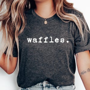 May include: A dark gray t-shirt with the word "waffles" printed in white on the front. The text is in a typewriter font.