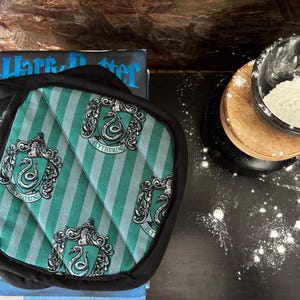 May include: A pair of green and white striped oven mitts with the Slytherin crest, resting on a book titled "Harry Potter". A measuring cup with flour sits on a wooden coaster, with flour sprinkled on the dark surface.