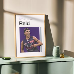 May include: A framed poster with the name "Reid" in black text. The poster shows a person in a sports uniform with their tongue out. The frame is light brown, and the poster is on a light green cabinet. There is a green vase and a small amber glass.