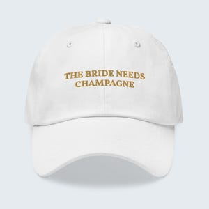 May include: White baseball cap with the embroidered text "THE BRIDE NEEDS CHAMPAGNE" in gold. The cap has a curved brim and an adjustable strap at the back. A fun accessory for a bachelorette party or bridal event.