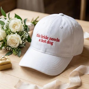 Może przedstawiać: Biała czapka z daszkiem z czerwonym napisem "this bride needs a hot dog". Czapka leży na drewnianej powierzchni, obok bukietu białych róż i pudełka na pierścionek.