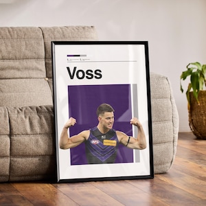 May include: Framed poster with the name "Voss" in white text above an image of a man flexing his biceps. The man is wearing a purple and navy blue athletic jersey. The poster is propped against a gray sofa. The poster is in a black frame.