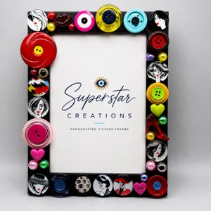 May include: A black rectangular picture frame decorated with various colorful buttons, beads, and circular illustrations of faces. The center white insert features the text Superstar CREATIONS HANDCRAFTED PICTURE FRAMES and an eye icon.