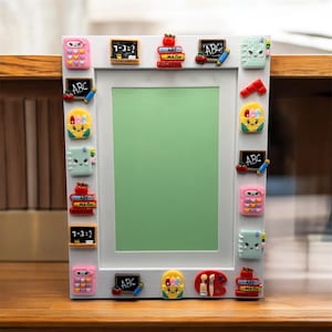 Puede incluir: Marco de fotos blanco decorado con adornos coloridos con temática escolar. El marco presenta diseños en miniatura de libros, manzanas, calculadoras y pizarras con las letras "ABC" y símbolos matemáticos. El marco tiene un centro verde.