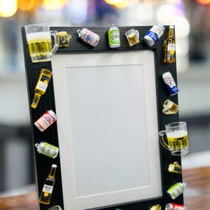 May include: A black rectangular picture frame decorated with miniature beer-themed ornaments. The frame features various small replicas of beer bottles, cans, and glass mugs filled with yellow liquid, arranged along the outer border.