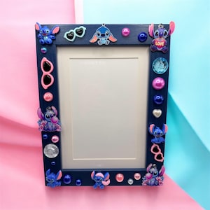 May include: A navy blue picture frame decorated with Stitch character embellishments, sunglasses, and pink and blue accents. The frame has a white interior and is adorned with various decorative elements.
