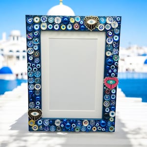 May include: A rectangular blue picture frame decorated with various blue and white eye-shaped beads and heart-shaped charms. The frame has a white interior and is set against a backdrop of white buildings and a blue sky.