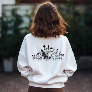 May include: White sweatshirt with a black floral design across the upper back. The design features silhouettes of various wildflowers and ferns. The sweatshirt is a casual, long-sleeved garment.