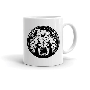 May include: White ceramic mug with a black and white illustration of two witches stirring a cauldron in a forest setting. The illustration includes ravens, stars, and a full moon.