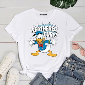 May include: White t-shirt featuring a graphic of Donald Duck in a blue sailor suit, red bow tie, and sailor hat. The text "FEATHERED FURY!" is displayed above the character in a blue burst. The shirt is paired with jeans and white sneakers.