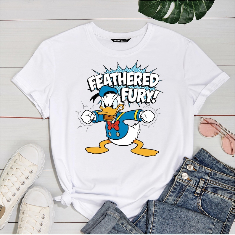May include: White t-shirt featuring a graphic of Donald Duck in a blue sailor suit, red bow tie, and sailor hat. The text "FEATHERED FURY!" is displayed above the character in a blue burst. The shirt is paired with jeans and white sneakers.