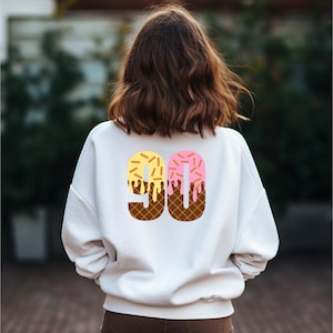 May include: White sweatshirt with a graphic of the number 90 made to look like an ice cream cone with yellow and pink ice cream and chocolate sprinkles.