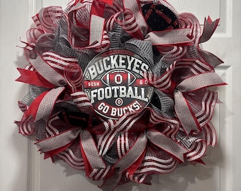 May include: A decorative wreath featuring red, gray, and white mesh ribbons. A circular sign in the center reads "Buckeyes Football Go Bucks" with a football graphic. The wreath is hanging on a white door.