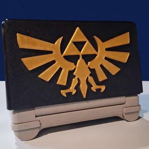 Puede incluir: Una funda negra para consola de juegos portátil con un emblema dorado con un diseño de triángulo y alas. La funda descansa sobre una base gris claro, con un fondo azul. Es un accesorio protector para un dispositivo de juego.